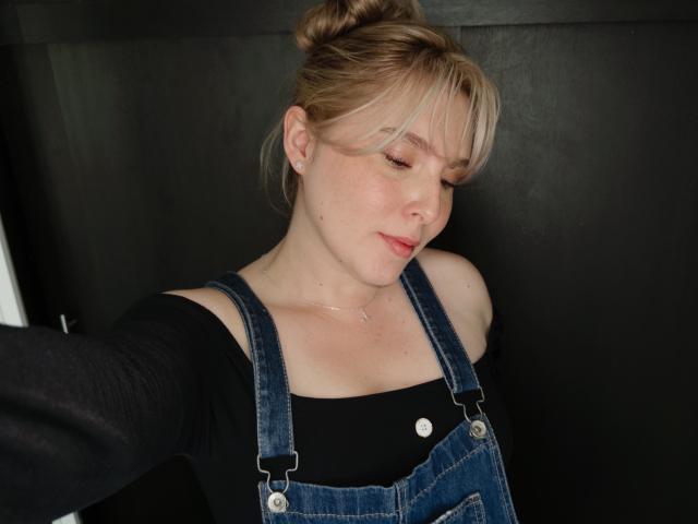 LindaDiamond with blonde hair in a bun, wearing denim overalls and a black top, eyes closed, slight smile, small breasts, fair skin, black background, selfie, casual.