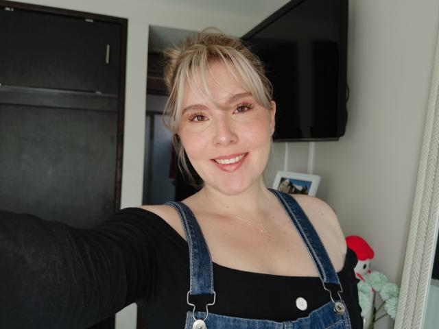 LindaDiamond with blonde hair, smiling, wearing denim overalls and a black shirt, showing a small nose and medium tits, holding out her arm for a selfie.