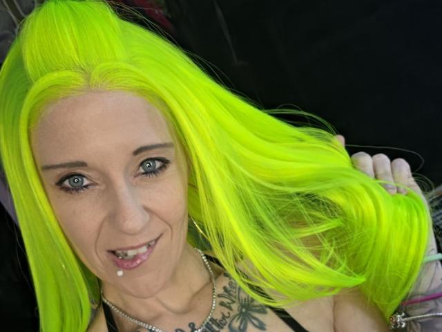 PebsStayThnx with bright neon yellow hair, blue eyes, and a tongue piercing, smiling at the camera, holding her hair. She's got a tattoo on her chest.