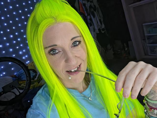 PebsStayThnx with neon yellow hair, blue eyes, and a tooth ring, holding a straw in her mouth, smiling. She's wearing a blue shirt with a necklace.
