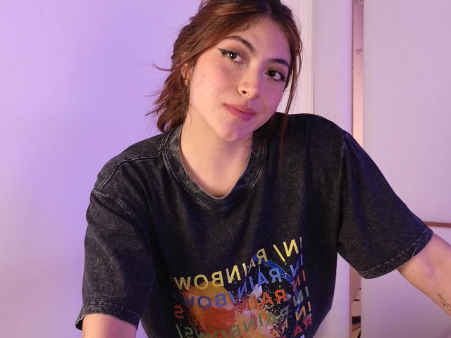 SpencerLe has light skin and brown hair, styled in a loose, slightly messy ponytail. She's wearing a dark, graphic T-shirt with colorful text and images. Her makeup includes bold, winged eyeliner. She has a small, visible nose piercing on her right nostril. Her expression is neutral, slightly smiling. Her arms are slightly out of frame, but a small tattoo is visible on her left forearm. The lighting is soft, with a purple hue. The background is simple and unobtrusive.