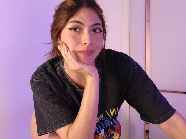SpencerLe, a young woman with light brown hair, is leaning forward, resting her chin on her hand. She has a slight smile, with dark, defined eyebrows and long, black eyeliner. She wears a dark, short-sleeved, graphic t-shirt with colorful designs. Her right hand, with a small tattoo on the wrist, supports her head. The light pink background and door frame are visible. The image has a soft purple and white light.