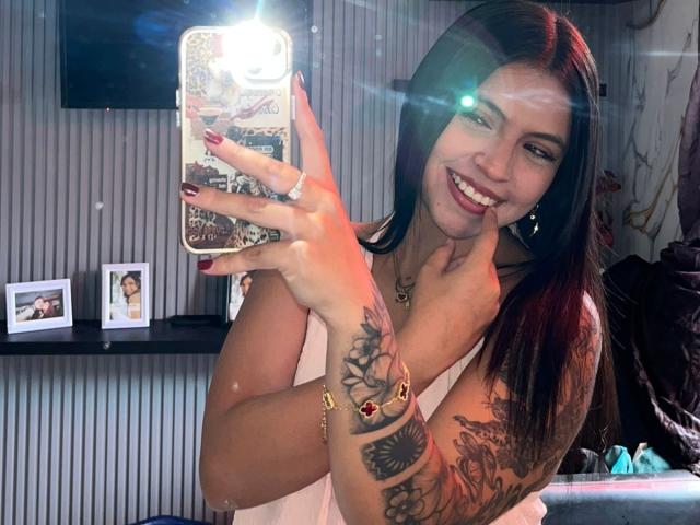 NyraVanel with long black hair, smiling, holding phone in front of mirror. She's showing off tattooed arm with colorful rings. Boobs visible under white top, red nails.