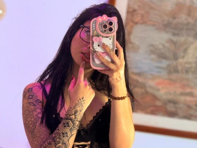 LunaDesire with black hair, glasses, tattoos on arms, pink lipstick, finger in mouth, holding pink Hello Kitty phone case, wearing black lace top, taking mirror selfie.