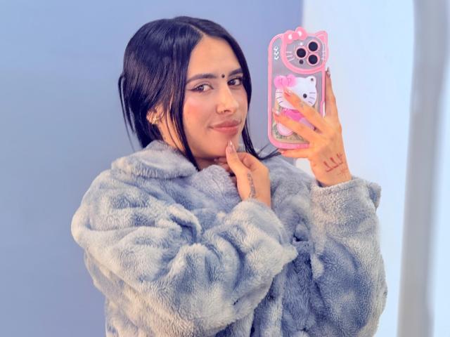 LunaDesire with dark hair, wearing a fluffy grey coat, holding a pink phone case with a Hello Kitty design, taking a mirror selfie with a subtle smile.