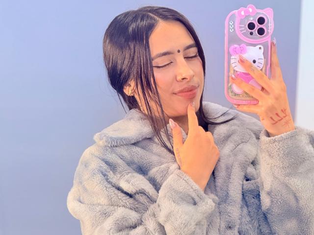 LunaDesire with dark hair, eyes closed, wearing a fluffy gray coat, holding a pink phone with a Hello Kitty case, index finger touching her lip, cute and relaxed.