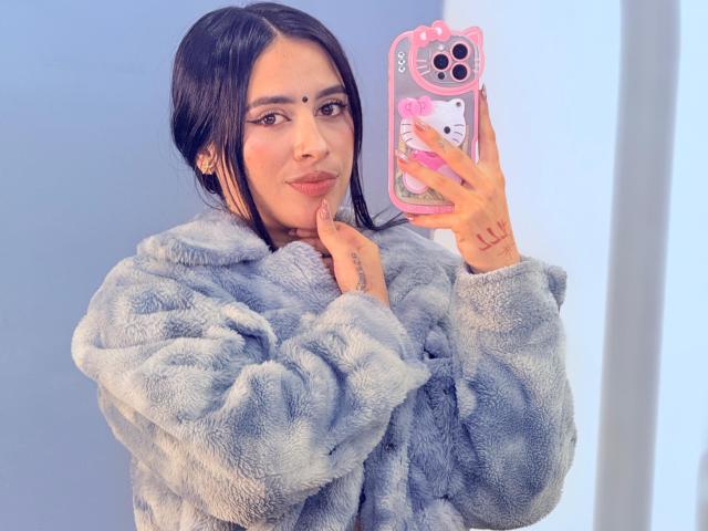 LunaDesire with dark hair, wearing a fluffy gray coat, holding a pink phone case with a Hello Kitty design, showing tattoos on her fingers and lips.