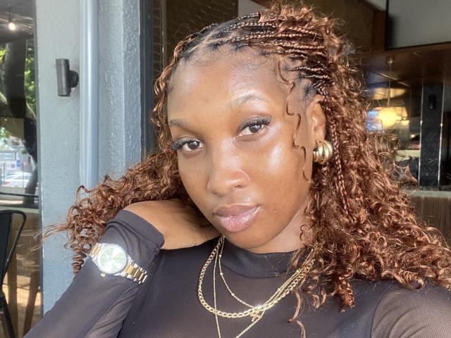 DarkCocoa has curly brown braids, a shiny dark skin, gold earrings, a black sheer top, a gold watch, and a gold necklace, looking at the camera with a neutral expression.
