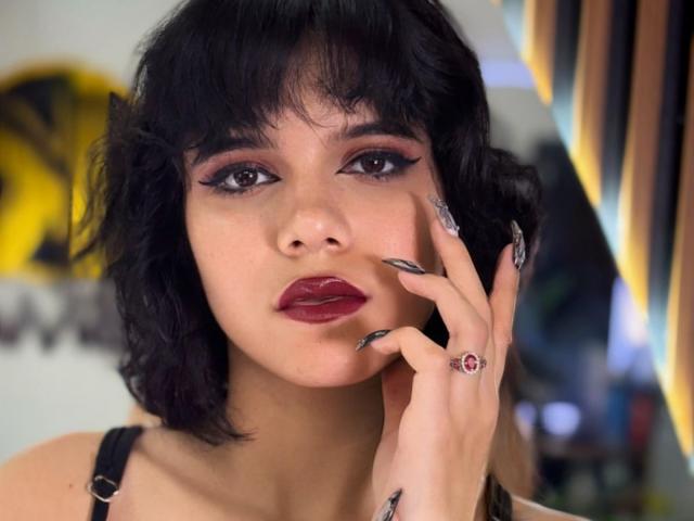RavenMorninsgstar with dark wavy hair, red lipstick, and dramatic eyeliner, touching her cheek with long, glittery nails, wearing a black strap top and a red ring.
