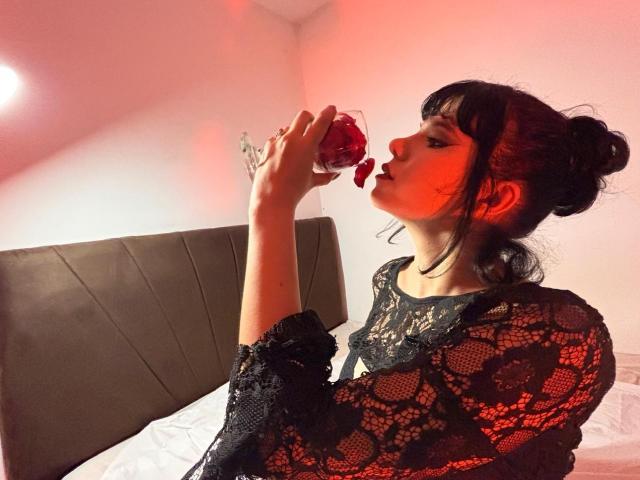 RavenMorningsstar with dark hair in a bun, wearing a black lace top, sips rose-petal wine, red light on her. She's got a cute face, small tits, and is sexy.