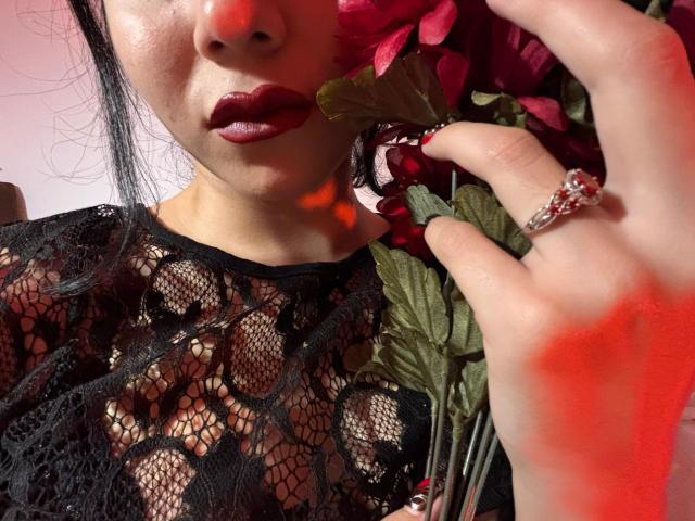 RavenMorninsgstar with red lipstick, holding a rose, wearing a black lace top, exposing her tits, ring on finger, red light casting shadow.