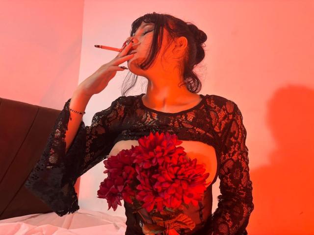 RavenMorninsgstar with black lace top, holding red flowers, smoking, tits visible, red light, black hair up, right hand to mouth, shadow on wall, bracelet on wrist.