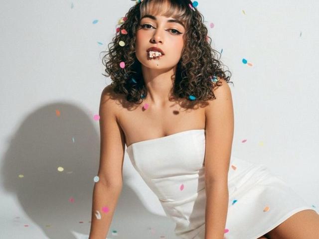MiaWhalker with curly brown hair, wearing a white strapless dress, has confetti on her face and body, with colorful confetti pieces falling around her, sitting on a white surface.