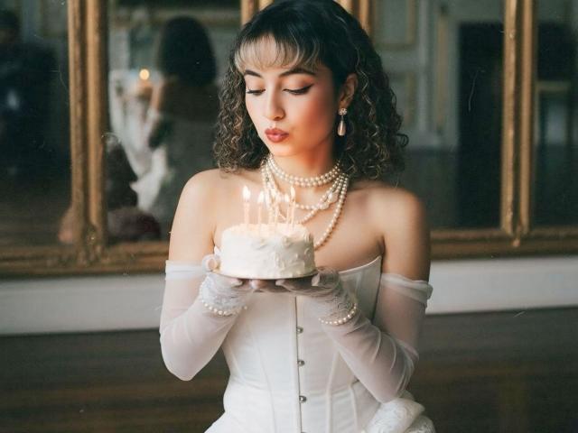 MiaWhalker with curly brown hair, white off-shoulder dress, pearl necklace, and gloves, blowing out candles on a small white cake, holding it with both hands, in a photo.