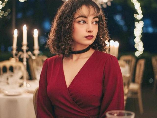 MiaWhalker with curly brown hair, wearing a red dress with a black choker, looking to the side with dark red lipstick. Boobs visible, candlelight in the background.