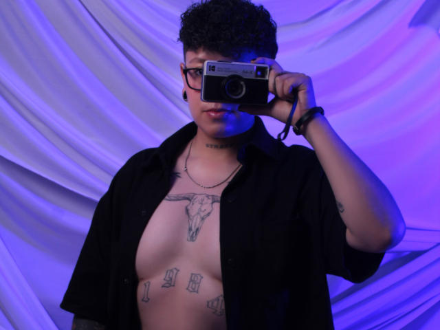 AllexNoa with curly black hair, glasses, and black shirt, holding a camera, showing off tattoos including a bull skull on chest and money symbols on stomach.