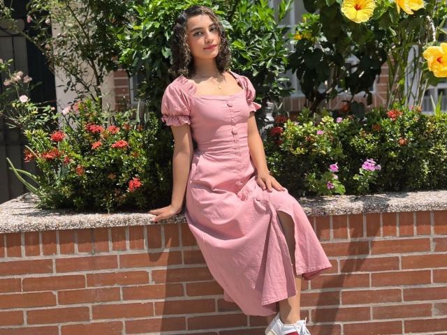 MiaWhalker sits on a brick wall, wearing a pink dress with buttons and a thigh slit, her ass slightly raised, curly brown hair, white sneakers, green plants behind.