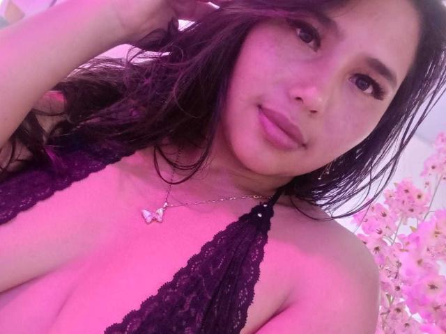 GemmaBellini has medium-length, wavy black hair and light brown skin. She wears a black lace halter top that reveals her cleavage. Her lips are slightly parted, and she has a relaxed, confident expression. She holds her hair with her right hand. A silver necklace with a butterfly pendant hangs around her neck. Pink and purple lighting casts a soft, warm glow over her face and upper body. Her eyes are slightly downcast, and her makeup is subtle. The image focuses on her upper body and face.