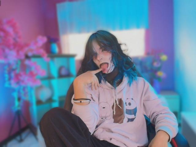 Dandii69 with blue-tipped hair, tongue out, licking finger, wearing a pink panda hoodie, black pants, sitting, phone on arm, pink and blue lighting, blurred background.