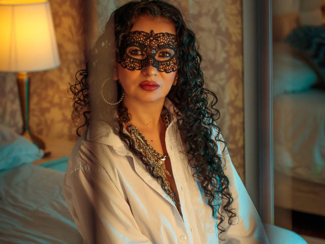 SamanthaAntolin has long, curly black hair and wears a detailed black lace mask with intricate patterns covering her eyes. She has dark red lipstick and large hoop earrings. Her white button-up shirt is slightly open, revealing a chunky gold necklace. The warm, yellowish lighting from a lamp on the left contrasts with the blueish light on her right. The background is a blurred bedroom. The image has a rich, textured feel with a mix of warm and cool colors.