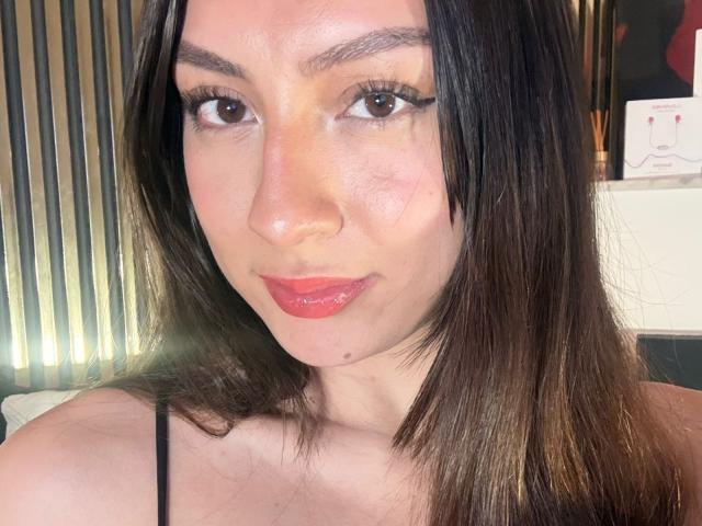 Luluberrie has shiny brown eyes, black eyeliner, red lipstick, and straight dark brown hair. Her skin is light with a slight blush. She's wearing a black strap.