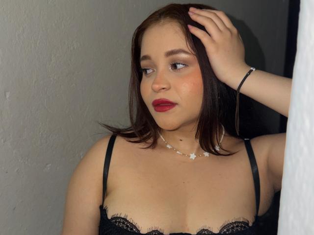 SamyMancini with brown hair, red lipstick, and black lace bra, hand in hair, wearing a star necklace, looking sideways, medium tits, light skin, slightly chubby, dark background.