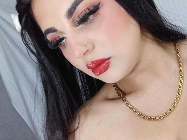 EmilyConore with glossy red lips, black eyeliner, and gold chain necklace, has black hair, a nose piercing, and a slight blush on her fair skin. Boobs not visible.