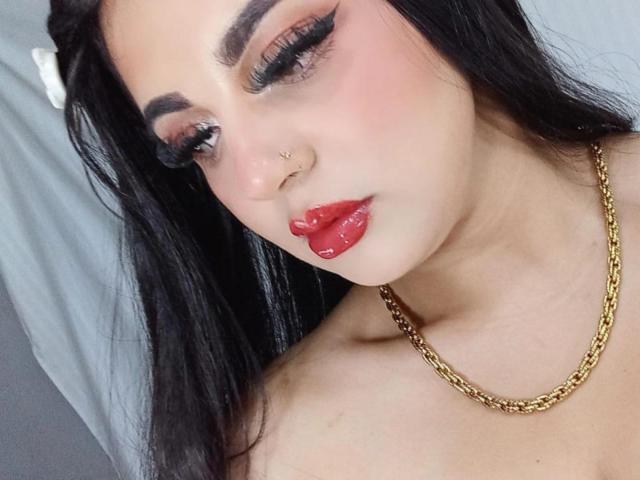 EmilyConore with black hair, red lips, gold chain necklace, nose ring, and thick eyelashes, looking down with slightly parted pink lips, showing off her tits in the photo.