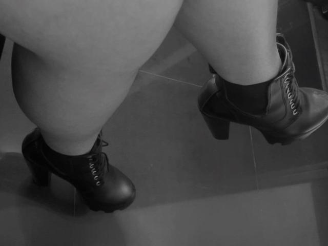 EmilyConore's chubby thighs and black, high-heeled, lace-up boots with chunky soles, standing on a dark, shiny floor, in a black-and-white photo.