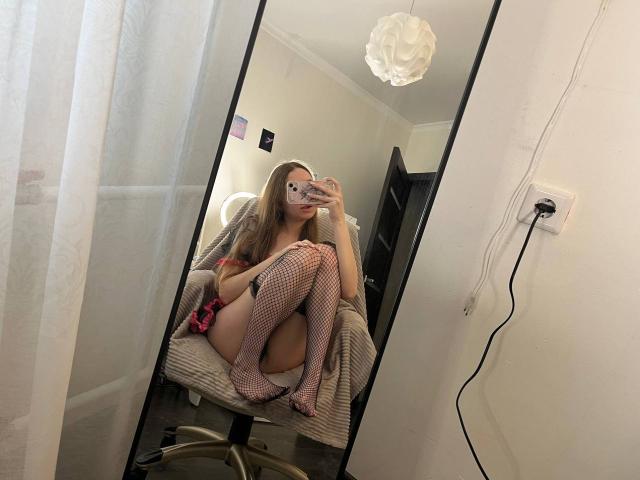 MaryanneSexy sitting in a chair, wearing fishnet stockings and pink lingerie, with her legs up, showing her pussy, and holding a phone in a mirror selfie.