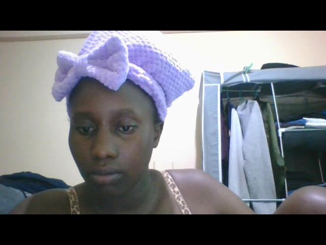 Tiiy has dark brown skin and a serious expression. She wears a purple crocheted headscarf with a large bow on top. Her spaghetti strap is beige. In the background, a metal shelf holds clothes, including a gray shirt. The image is a photograph with a soft, slightly blurry focus. Tiiy's head is slightly tilted to the right. Her eyes look to the left of the camera.