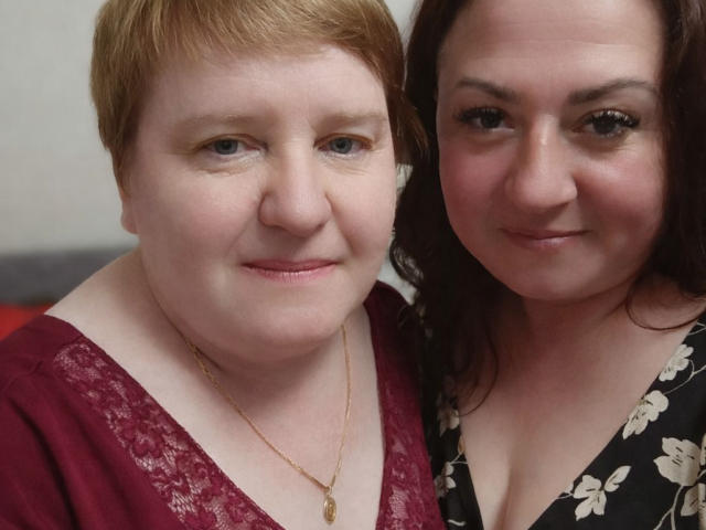 MollyAndJane with short red hair and pale skin, wearing a red lace top, stands next to MollyAndJane with brown hair and pale skin, in a black floral top.