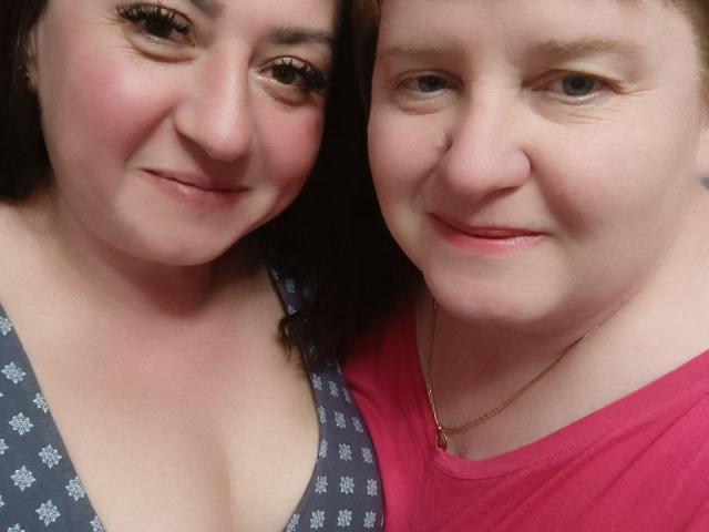 Close-up of MollyAndJane, two women with fair skin and short hair. MollyAndJane on the left has dark brown hair, thick eyebrows, and wears a gray patterned top with a low neckline. MollyAndJane on the right has lighter hair, thin eyebrows, and wears a pink shirt with a gold necklace. Both women have slight smiles and are looking at the camera. The image is a selfie with a focus on their faces.