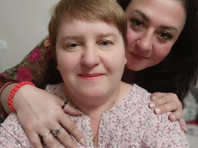 MollyAndJane with short blonde hair, pink floral shirt, and red bracelet, has her ass grabbed by MollyAndJane with dark hair, floral jacket, and red bead bracelet.