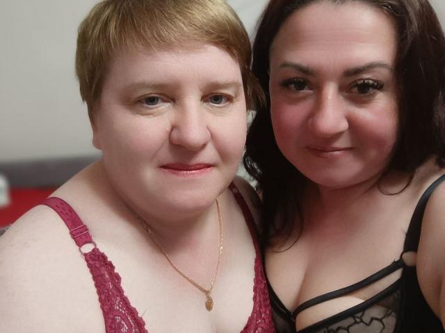 MollyAndJane with short red hair in a red lace bra and Jane with dark brown hair in black lace lingerie, both smiling at the camera, showing their tits.