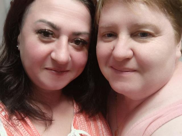 MollyAndJane has dark brown hair, medium-sized tits, and a pink dress. Jane has short blonde hair, pale skin, and small tits, wearing a pink top. Both smile.