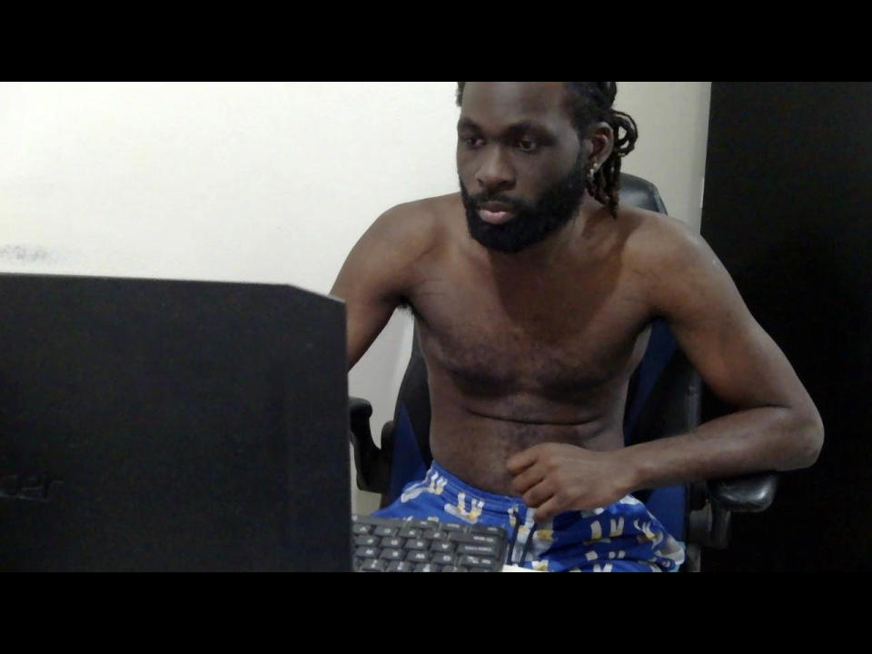 Um Darkkras sem camisa, com pele escura, dreadlocks e barba, vestindo shorts azuis com padrões brancos, sentado em uma mesa, olhando para um computador com um teclado na frente..