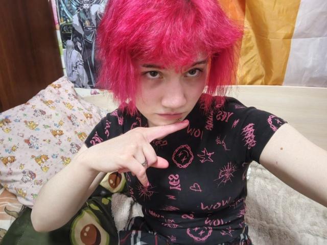 Katlyn Renteria mit leuchtend rosa, zerzaustem Haar, trägt ein schwarzes Oberteil mit rosa Mustern und zeigt mit einem Finger auf ihre leicht geöffneten Lippen. Sie sitzt auf einem Bett mit bunten Kissen..