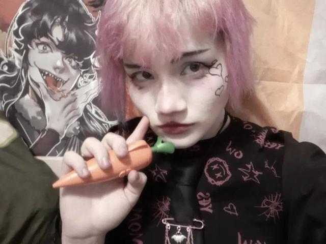 KatlynRenteria, com cabelo rosa, segurando uma cenoura laranja, vestindo uma blusa preta com desenhos rosa, tem tatuagens de coração no rosto, olhando para a câmera, com um pôster de vampiro..