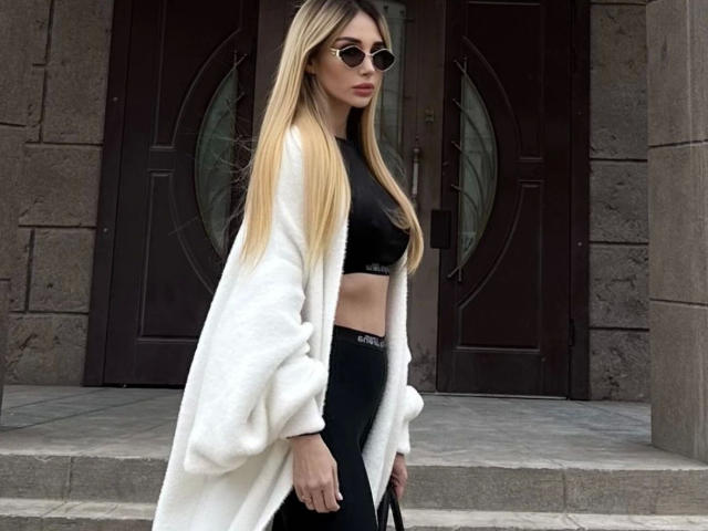 PlayfullBella-hot stands in front of a dark brown door with circular windows. She has long, straight blonde hair and wears round sunglasses. She's dressed in a black sports bra, black leggings with "Puma" text, and a white, fluffy coat. Her hands hold a black bag. The stone building has a textured, gray exterior. The composition focuses on her from the side, highlighting her casual yet stylish outfit.
