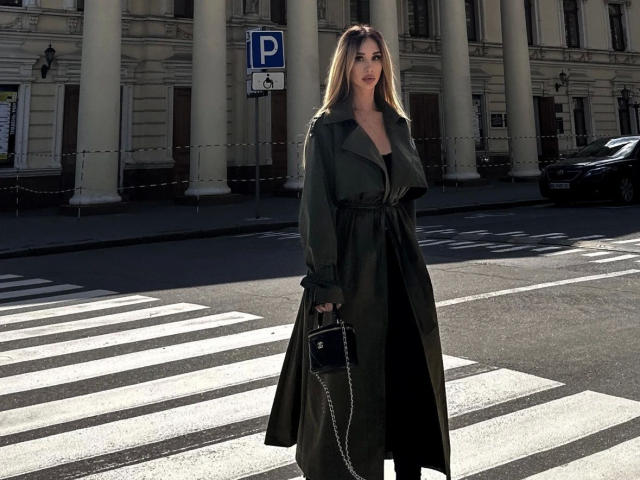 PlayfullBella-hot stands in a crosswalk, wearing a long, dark green coat with a belt, and a black crossbody purse with a chain. She has long blonde hair and red lipstick. The background includes a building with tall columns and a "P" parking sign. The image is well-lit with shadows highlighting the crosswalk stripes. Her confident stance contrasts with the muted, classical architecture.