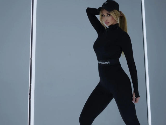 PlayfullBella-hot with blonde hair, wearing a black long-sleeve bodysuit, black cap, and tight pants, standing with one hand on her head, showing her ass.