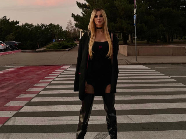 PlayfullBella-hot stands in a crosswalk, front and center, with a dark, black dress, thigh-high shiny black boots, and a black jacket. Her long blonde hair falls straight over her shoulders. She wears a black choker and has a small red emblem on her dress. The crosswalk has white and red stripes, with a tree-lined background and a street. The image has a muted, evening light. Her confident, slightly relaxed posture contrasts with the structured, striped crosswalk.