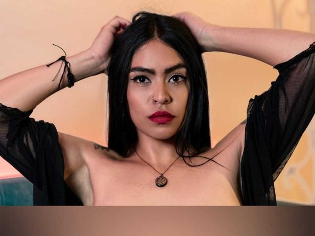 StellaBeckker with black hair, arms raised, wearing a sheer black top, red lipstick, nose ring, and black bracelet, showing her tits, with a pendant necklace.