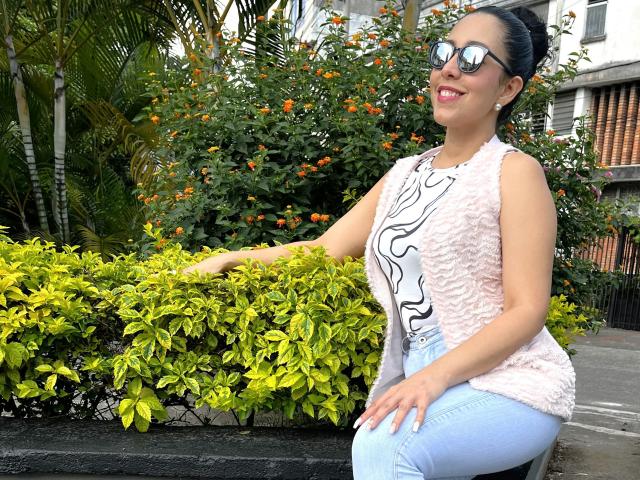 KattyRivera with big tits, wearing a pink fuzzy vest, white and black patterned top, blue jeans, and reflective sunglasses, leaning on green shrubbery, smiling, with orange flowers behind.