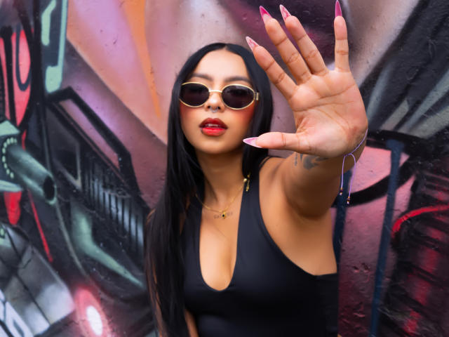 ChloeMoretti stands in front of a graffiti-covered wall, reaching out with her right hand. She has long black hair, wears sunglasses, and sports a black sleeveless top with a deep V-neck. Her nails are long and painted pink. Her lips are red and she wears a small necklace. The graffiti includes bold colors like pink, green, and black. Her tattoo is visible on her right wrist. The image is sharp with a focus on her hand.