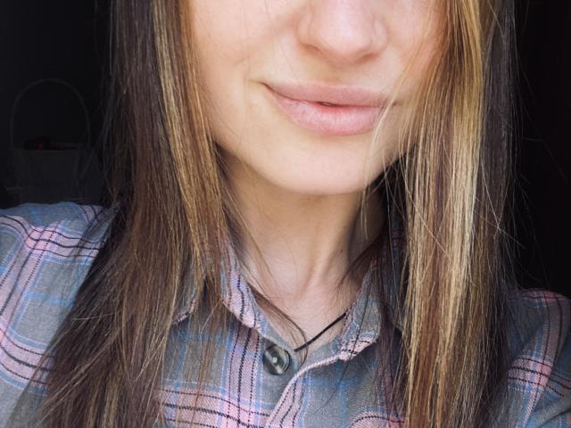 Close-up of Milagres' face, showing her lips slightly parted with a subtle smile. Her long brown hair has blonde highlights, with strands falling over her face. She wears a plaid shirt with pink, blue, and gray lines. A black cord necklace is visible. The image focuses on her lower face and hair, with soft, natural lighting highlighting her skin and hair. The background is blurred and dark.