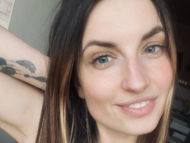 Close-up of Milagres with fair skin, blue eyes, and brown hair with blonde highlights. She has thick, dark eyebrows and is smiling with slightly parted lips. Her right arm is raised, showing a black tattoo with a feather design on her inner bicep. Her long hair is slightly messy. The light is soft and natural, highlighting her smooth skin. The composition is tight, focusing on her face and upper torso. She has a relaxed, confident expression.