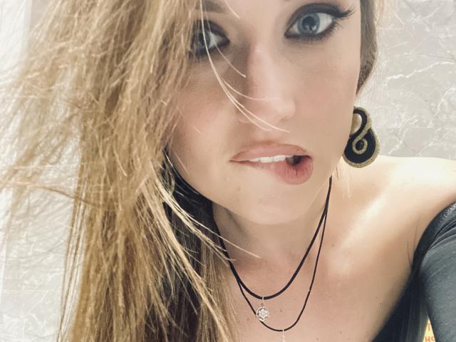 Milagres has blonde hair, blue eyes, and is wearing black eyeliner with long lashes. She has small tits, a black off-shoulder top, and black ear gauges.