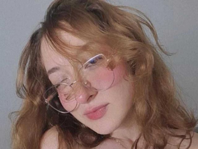 CattalinaBotero with wavy brown hair, pink-tinted glasses, and pink flushed cheeks, looking slightly to the side with a subtle smile, showing her soft pink lips.